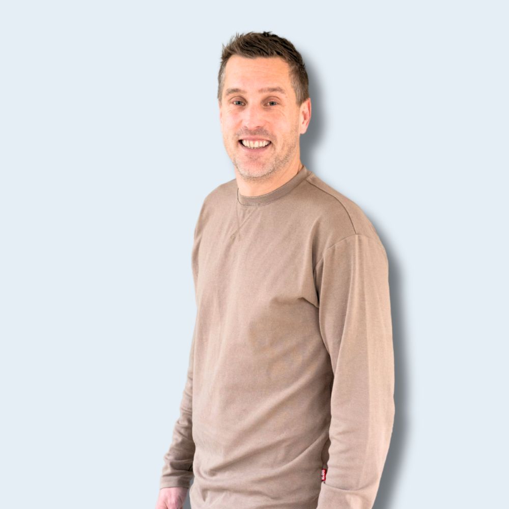Man die relaxt staat en glimlacht naar de camera. Hij draagt een beige lange mouwen t-shirt en licht blauwe jeans. 