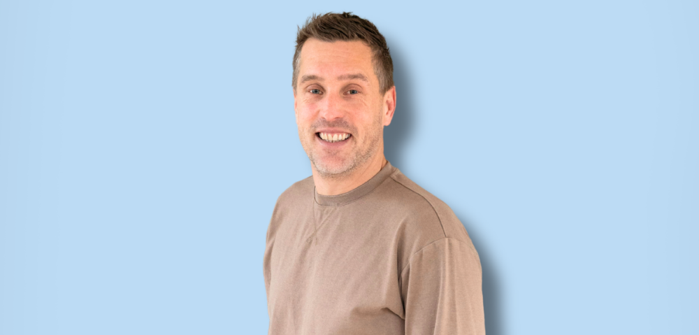 Man die relaxt staat en glimlacht naar de camera. Hij draagt een beige lange mouwen t-shirt en licht blauwe jeans. 
