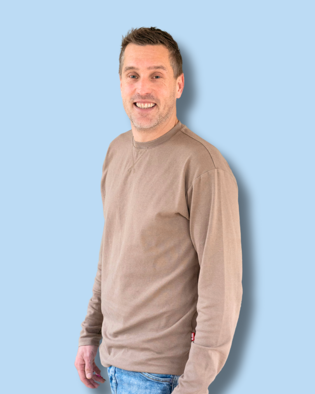 Man die relaxt staat en glimlacht naar de camera. Hij draagt een beige lange mouwen t-shirt en licht blauwe jeans. 